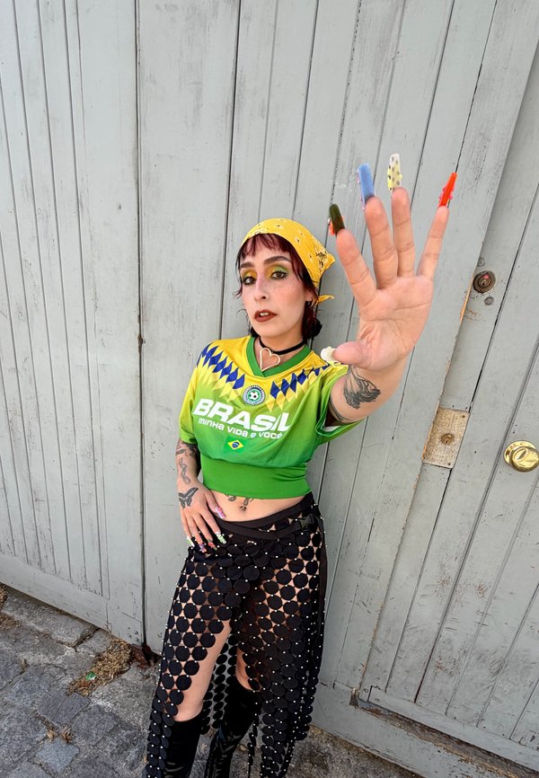 Persoon draagt een gele bandana, een Braziliaans sportshirt en een zwarte netrok met lange gekleurde nagels, houdt de hand richting de camera tegen een houten deur.
