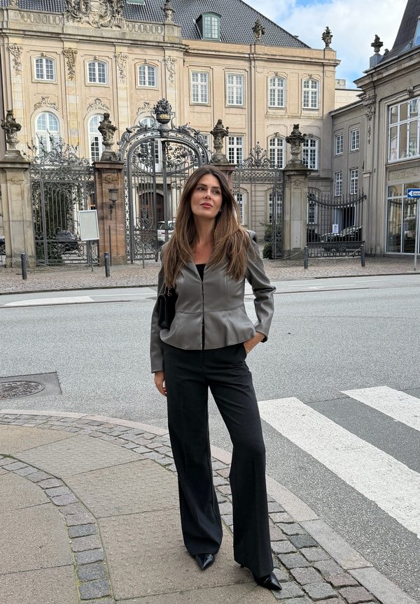 Woman with long brown hair wearing gray jacket and black pants stands on sidewalk near a historic building with ornate iron gates.