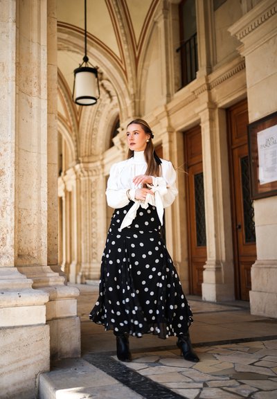 Mujer con blusa blanca y falda negra con lunares está de pie bajo un pasillo arqueado de piedra con puertas de madera y luz colgante.