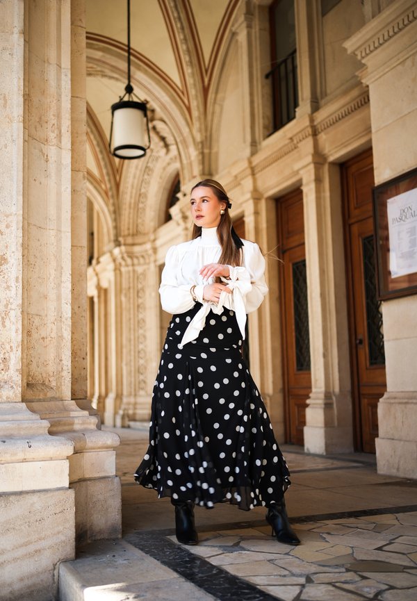 Kvinde iført hvid bluse og sort nederdel med prikker står under en hvælvet stenkorridor med trædøre og hængende lys.