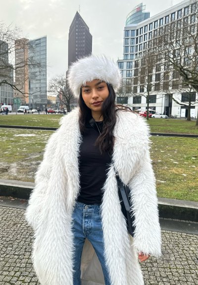 Mujer con abrigo blanco de peluche y gorro a juego, camiseta negra, jeans azules, de pie en un parque urbano con edificios modernos y árboles sin hojas.