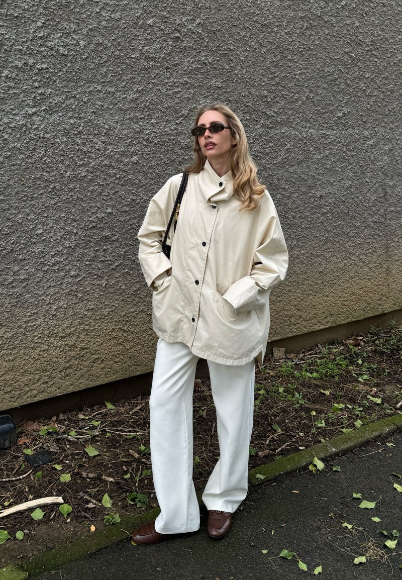Woman wearing beige oversized buttoned jacket, white pants, brown shoes, and black sunglasses standing by textured gray wall outdoors.