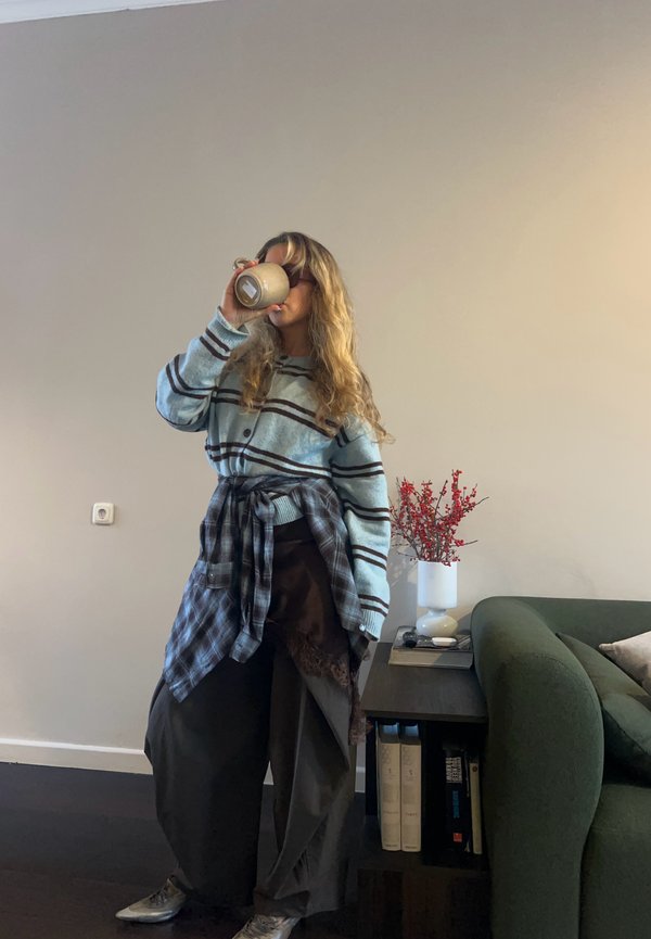 Blue striped cardigan over a brown skirt, paired with oversized pants. Holding a beige mug, standing near a green sofa and white flowers.