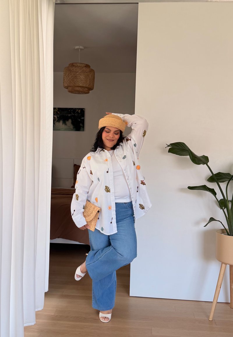 Femme portant une chemise blanche à motifs, un jean bleu, un chapeau beige et des sandales blanches, posant dans une pièce avec un sol en bois et une plante en pot.