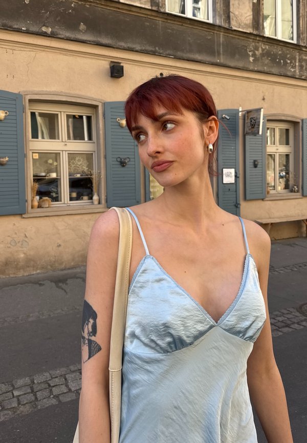 Giovane donna con capelli rossi e orecchini, indossa un vestito leggero azzurro e una borsa a tracolla, in piedi su una strada con finestre con persiane blu dietro di lei.