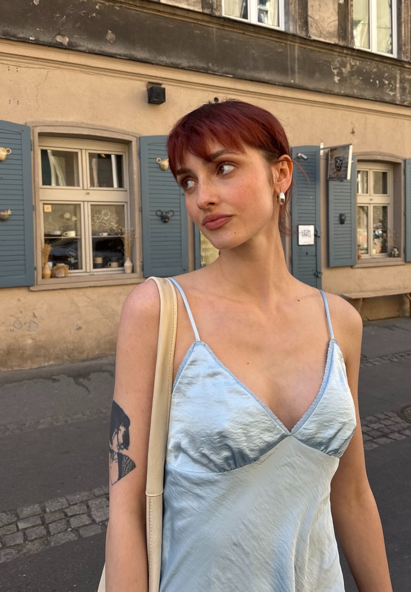 Jeune femme aux cheveux roux et boucles d'oreilles, portant une robe nuisette bleu clair et un sac cabas, debout dans une rue avec des fenêtres à volets bleus derrière elle.
