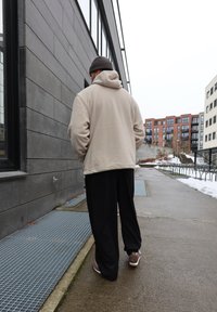 Άνδρας με μπεζ φούτερ με κουκούλα και μαύρο παντελόνι περπατάει σε αστικό πεζοδρόμιο δίπλα σε γκρίζο κτίριο με χιονισμένες περιοχές στο έδαφος.