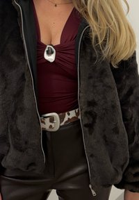 Femme portant un haut froncé bordeaux avec un pendentif en forme de coquillage, une veste noire en fausse fourrure, une ceinture imprimée vache et un pantalon en cuir noir, aux longs cheveux blonds.