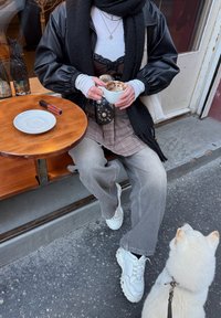 Veste bomber noire, jupe à carreaux, chemise blanche à manches longues, jean gris, baskets blanches. Tient une tasse de café ; un petit chien blanc à proximité. Table en bois.