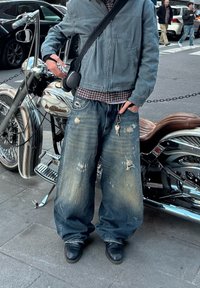 Casaco de denim azul claro, camisa xadrez, calças largas desbotadas com buracos desfiados, botas pretas e uma motocicleta ao fundo.