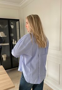 Camicia oversize a righe blu e bianche con orlo curvo, dettagli con bottoni sul retro e maniche ample. Indossata con jeans a vita alta.