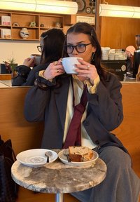 Femme portant des lunettes et un blazer, buvant du café dans un café, assise à une table avec une assiette de gâteau et des couverts.