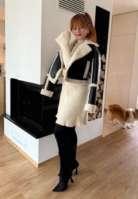 Γυναίκα με κόκκινα μαλλιά που φοράει μαύρο και μπεζ μπουφάν με επένδυση shearling, μπεζ πλεκτό φόρεμα και μαύρα ψηλοτάκουνα μπότες, στέκεται κοντά στο τζάκι με ένα σκύλο δίπλα της.