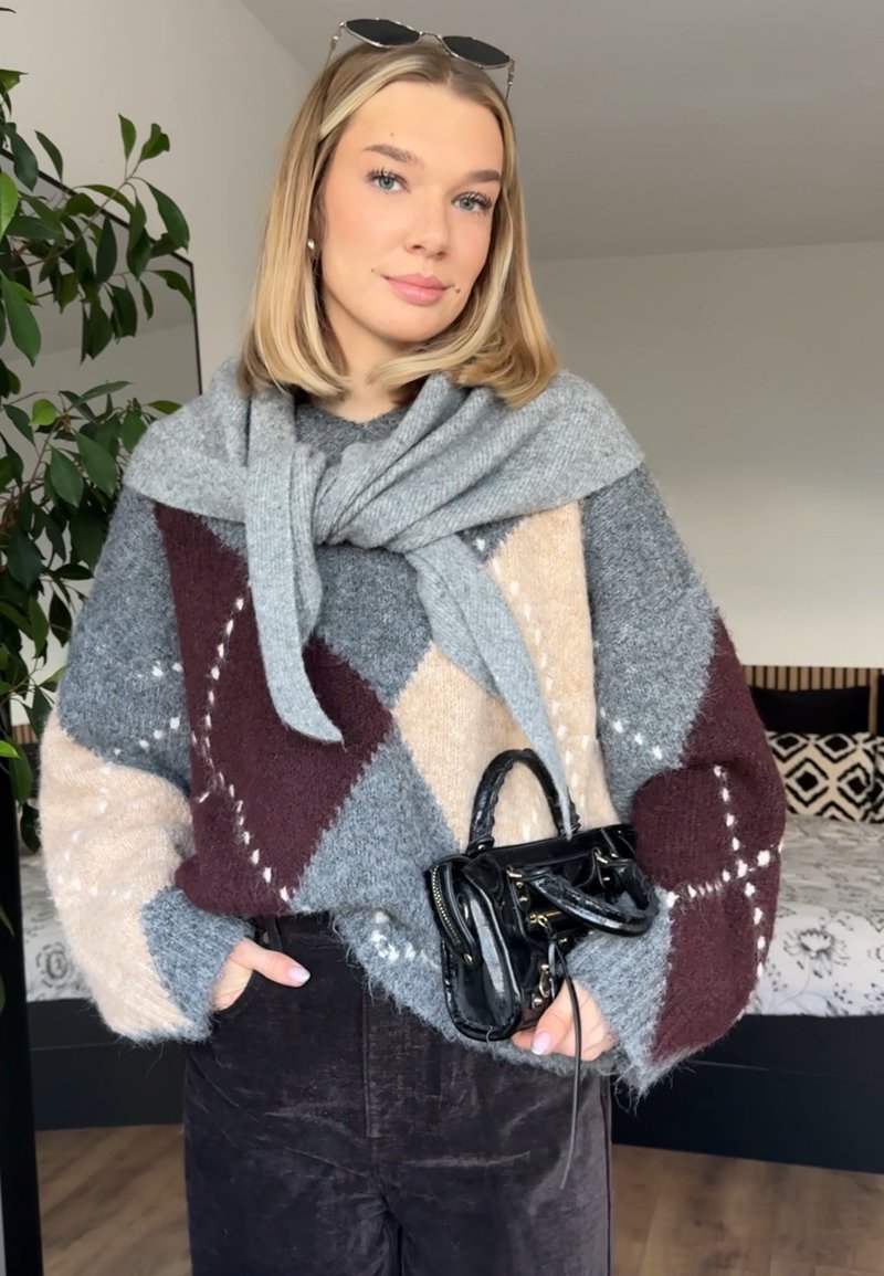 Jeune femme portant un pull à motifs gris, beige et bordeaux avec une écharpe grise, tenant un petit sac à main noir à l'intérieur près d'un lit.