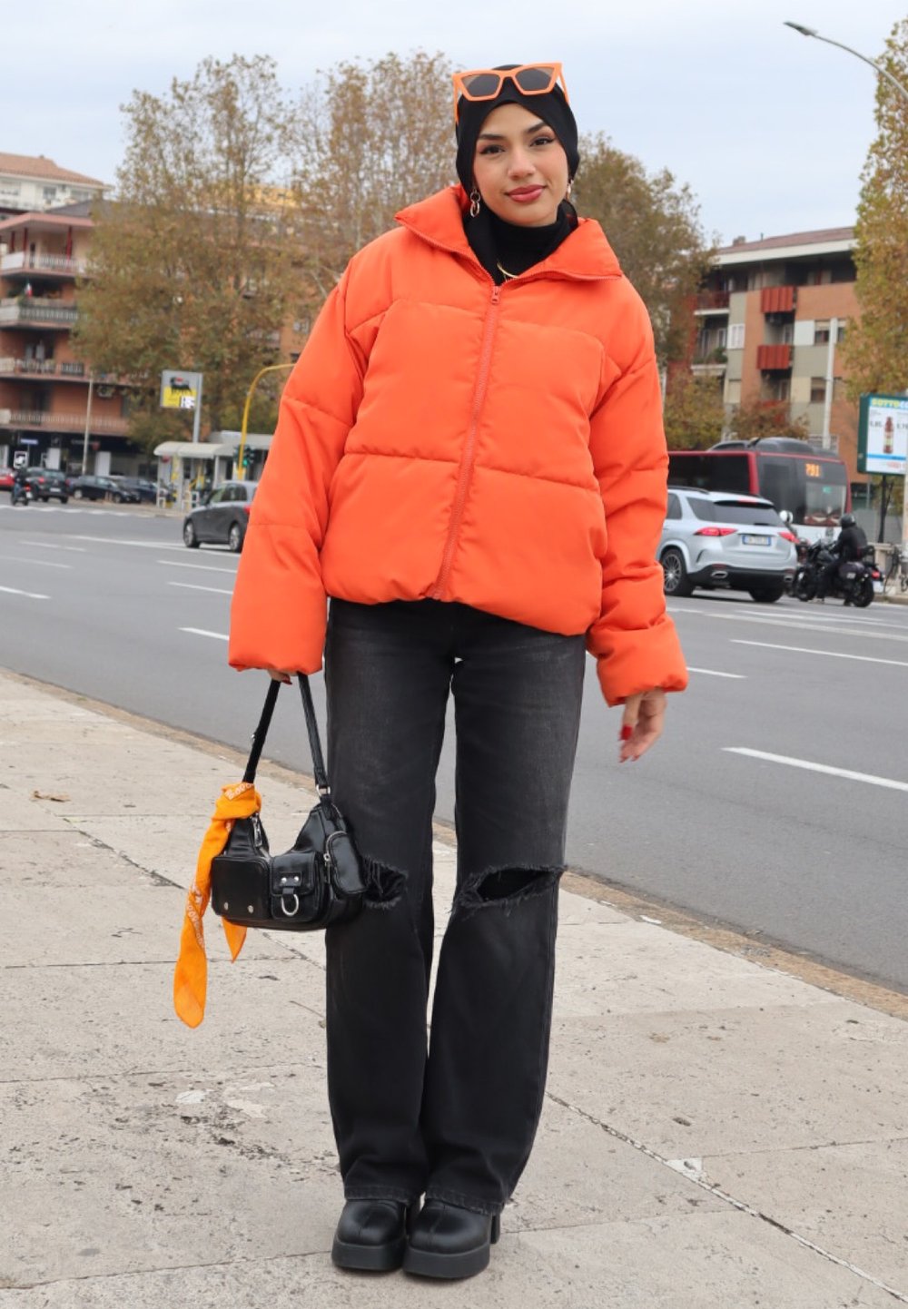 Veste rembourrée orange vif avec de grandes sections matelassées. Jean noir déchiré et chaussures à plateforme noires et épaisses. Petit sac à main noir avec un accent orange.