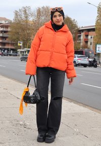 Veste rembourrée orange vif avec de grandes sections matelassées. Jean noir déchiré et chaussures à plateforme noires et épaisses. Petit sac à main noir avec un accent orange.