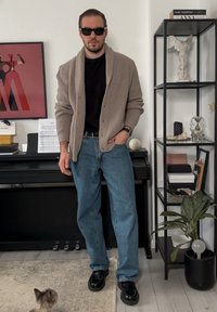 Cardigan côtelé beige par-dessus une chemise noire, jean bleu et chaussures noires. Accessoires : lunettes de soleil et une montre. Cadre intérieur avec décoration.