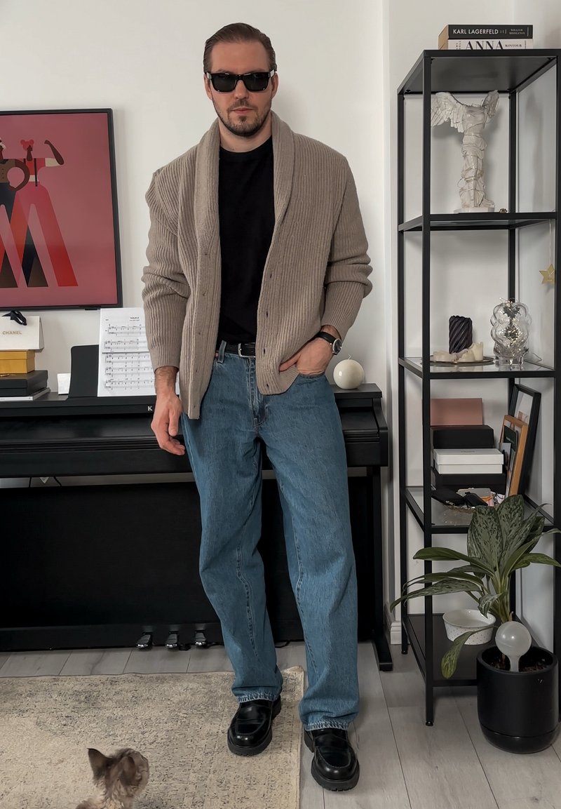 Cardigan beige côtelé porté sur une chemise noire, jean bleu, et chaussures noires. Accessoires : lunettes de soleil et montre. Cadre intérieur avec décoration.