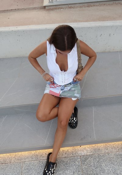 Top blanco sin mangas con un escote en V profundo y detalle de atado en la parte delantera, combinado con una minifalda de mezclilla rasgada y zapatos negros de slip-on con embellecimientos.