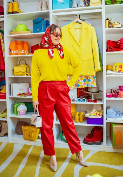 Mujer con top amarillo, pantalones rojos, bufanda roja y gafas de sol está delante de ropa y accesorios coloridos en estantes.