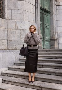 Przesadnie duży szary sweter dziany, przewiązany w talii, zestawiony z czarną skórzaną spódnicą midi i brązowymi czółenkami z migdałowym noskiem. Ciemna torebka na ramieniu.