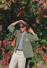 Giacca di denim verde oliva con maniche oversize, maglietta grigia e jeans bianchi; borsa intrecciata marrone e occhiali da sole scuri. Sfondo di foglie autunnali colorate.