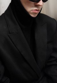 Blazer noir aux épaules structurées, col montant, accompagné d'un col roulé assorti. Lunettes avec verres foncés. Texture de tissu lisse et mate.