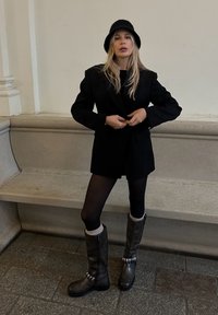 Černý oversized blejzr, černý top, černé punčochy, hnědé kozačky ke kolenu se stříbrnými detaily a černý klobouk bucket. Jednoduché, jednobarevné barevné schéma.