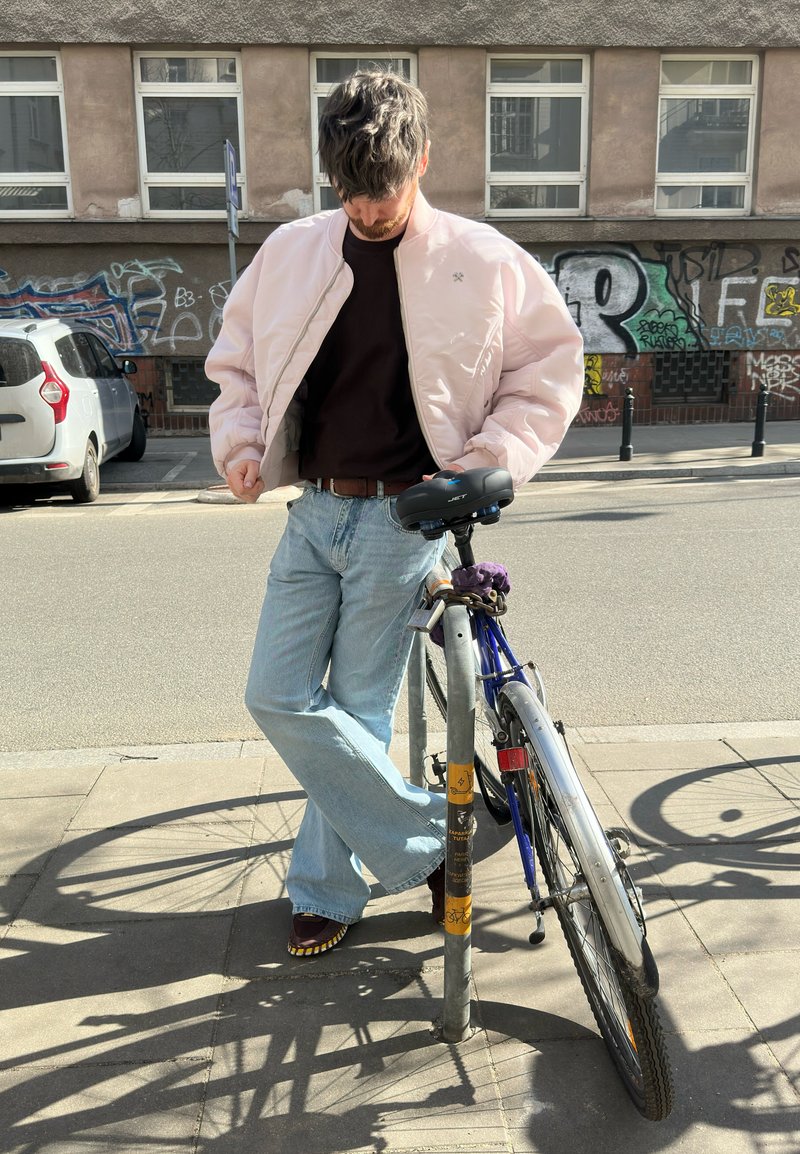 Mann in hellrosa Jacke und Jeans lehnt an einem verschlossenen Fahrrad auf einem sonnigen städtischen Bürgersteig mit Graffiti an einem Gebäude im Hintergrund.