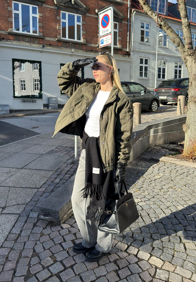 Femme se protégeant les yeux du soleil, portant une veste verte, des gants noirs, un pantalon gris, tenant un sac à main noir, debout sur une rue pavée près de voitures garées.