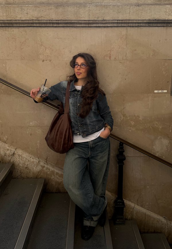 Kvinde i denimjakke og jeans læner sig på gelænderet ved trappen, holder en iskaffe og har en brun taske over skulderen mod en beige stenvæg.