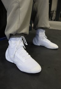 Person wearing white high-top trainers and grey joggers standing on a black textured floor.