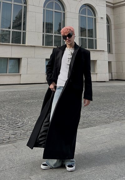 Persona con cabello rosa que lleva un abrigo largo negro, camisa blanca, jeans azul claro, gafas de sol negras y zapatillas de deporte, de pie en una calle adoquinada.