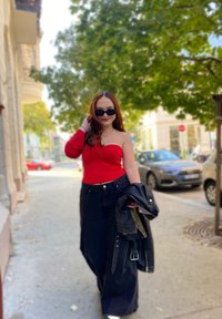 Κόκκινο τοπ με ένα ώμο, μαύρα τζιν ψηλής μέσης με φαρδιά πόδια, μαύρο δερμάτινο μπουφάν και γυαλιά ηλίου, περπατώντας σε έναν δρόμο με δέντρα.