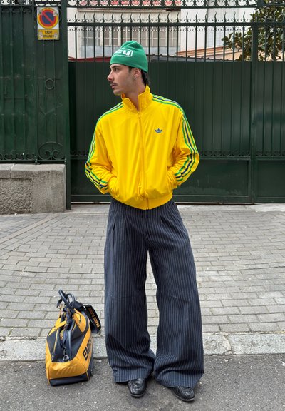 Hombre con chaqueta amarilla de Adidas, gorro verde y pantalones anchos de rayas finas está de pie en la calle junto a una bolsa amarilla y negra.