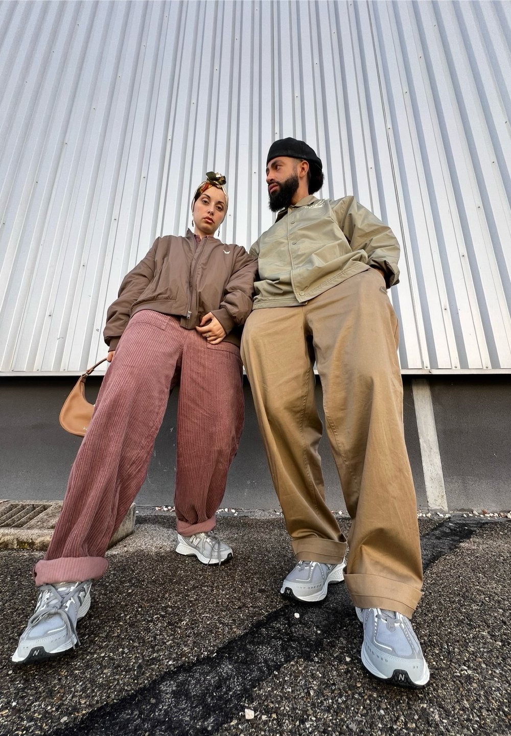 Δύο άτομα ντυμένα με casual ρούχα που στέκονται δίπλα-δίπλα. Το ένα φοράει ένα καφέ bomber jacket και ροζ παντελόνι από κοτλέ, ενώ το άλλο φοράει ένα ανοιχτό πράσινο μπουφάν και μπεζ παντελόνι. Και οι δύο έχουν γκρι sneakers.