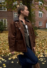 Smeđa zamšna jakna s kontrastnim crnim ovratnikom i patentnim zatvaračima, s prednjim džepovima, nosi se preko kariranog košulje i traperica.