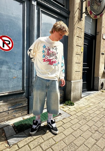 Camisa blanca de mangas largas con gráficos coloridos y estrellas, combinada con pantalones cortos de mezclilla azul claro y zapatillas negras. Entorno urbano con paredes texturizadas.