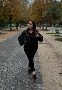 Eine Person in einer schwarzen Lederjacke geht auf einem Kopfsteinpflasterweg und hält eine kleine schwarze Tasche, mit herbstlichen Bäumen im Hintergrund.