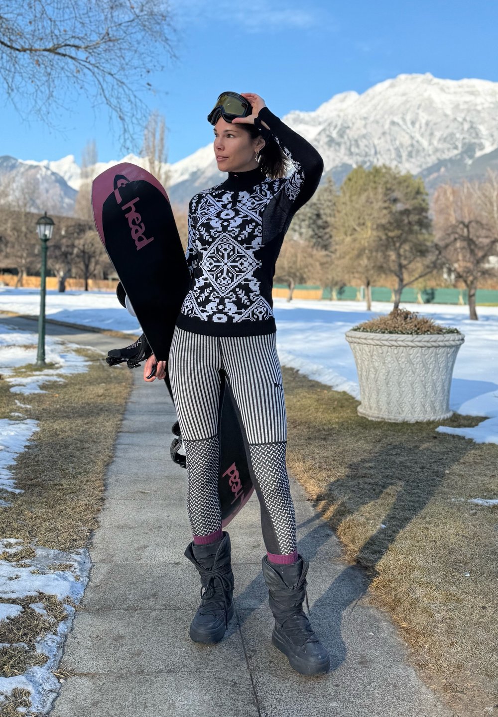Femme en tenue de sport d'hiver tenant un snowboard, portant des lunettes de protection sur la tête, debout sur un chemin enneigé avec des montagnes en arrière-plan.