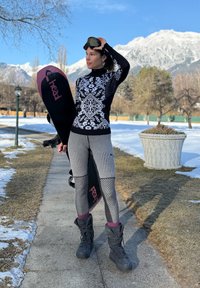 Femme en tenue de sport d'hiver tenant un snowboard, portant des lunettes de protection sur la tête, debout sur un chemin enneigé avec des montagnes en arrière-plan.