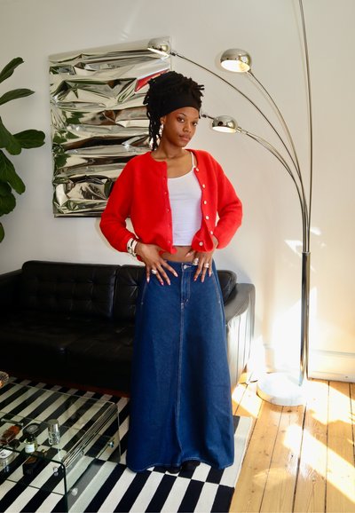 Suéter rojo de botones, hecho de lana suave; combinado con una falda de mezclilla larga con una abertura frontal y costuras contrastantes. Sofá negro al fondo.