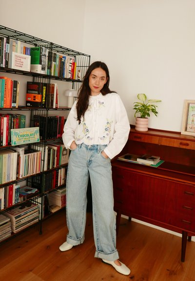 Blusa blanca bordada con detalle de lazo, jeans de pierna ancha de tiro alto en color azul claro y zapatos blancos tipo slip-on, con fondo de estantería.