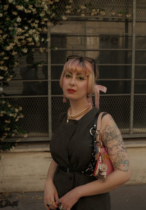 Donna con capelli corti tinti di rosa, trucco colorato e tatuaggio di gatto sul braccio, che indossa un vestito scuro e tiene una borsa con portachiavi.