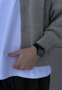 Montre noire avec cadran rond, accents argentés et bracelet en tissu texturé ; cardigan gris côtelé porté sur un t-shirt blanc ; main posée sur la taille.