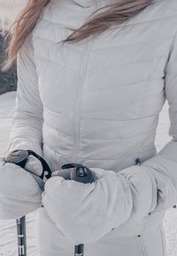 Personne portant une veste matelassée blanche et des moufles, tenant des bâtons de ski noirs et argentés dans un environnement enneigé en plein air.