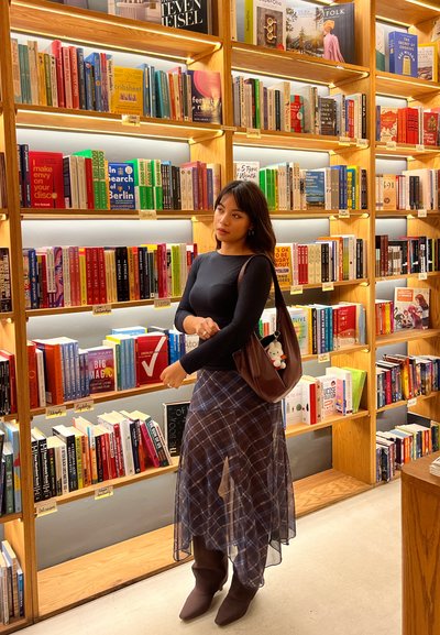 Una mujer con una blusa negra y una falda a cuadros está de pie frente a estanterías de madera bien iluminadas llenas de libros coloridos en una librería.