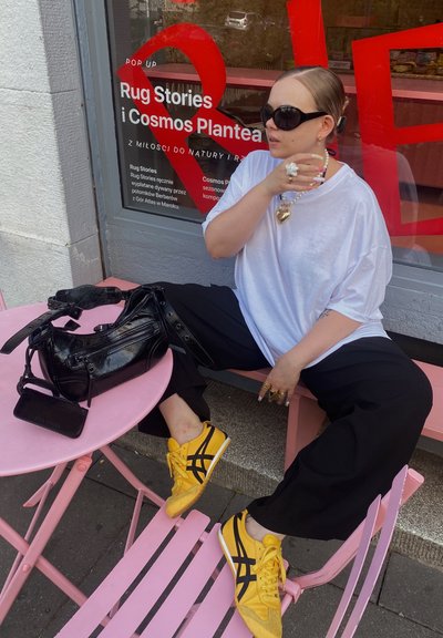 Bolso negro sobre una mesa rosa, individuo con camiseta blanca de gran tamaño y pantalones negros, usando zapatillas amarillas con rayas negras, sentado al aire libre.