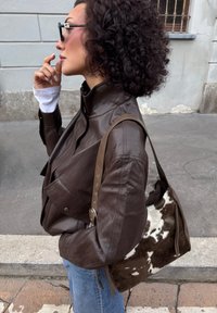 Brązowa skórzana kurtka z wysokim kołnierzem, w połączeniu z torbą w brązowo-białe wzory. Lekkie niebieskie dżinsy i czarne okulary dopełniają całość.