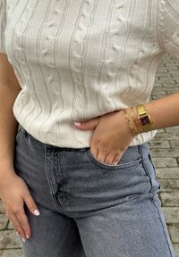 Pulôver curto de malha canelada creme, jeans cinzentos de cintura alta, relógio de pulso dourado com mostrador roxo e uma pulseira de corrente dourada.